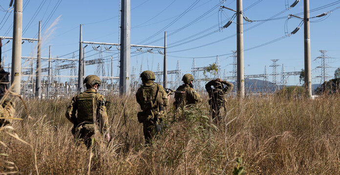 Army personnel move through long grass near an electricity substation