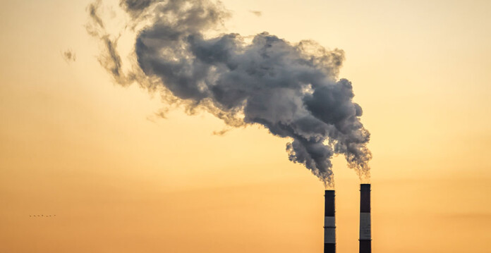 Smoke pours from chimney stacks at industrial factory with hazy orange sky overhead