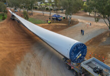 Locals get meet and greet with wind turbine blade Massive wind turbine blade on a truck bed with red dirt ground underneath it