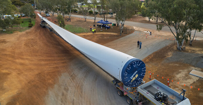 kings-rocks-community-event Massive wind turbine blade on a truck bed with red dirt ground underneath it