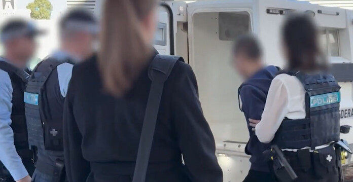 Police officers surround a police van while a handcuffed man with his face blurred is moved towards the vehicle