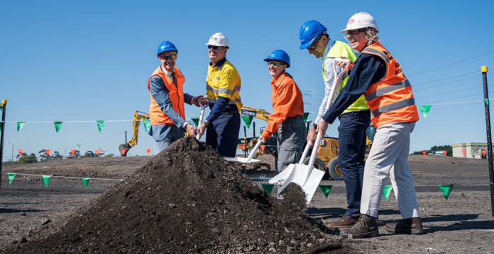Executives in high-vis vests and hard hats turn first sod at battery project site with blue sky overhead