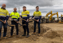 Flow Power unveils proposed Bennetts Creek Battery Stage 2 Four male workers in high-vis safety wear shovel dirt at battery project site with heavy plant in the background