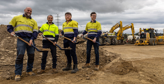 Four male workers in high-vis safety wear shovel dirt at battery project site with heavy plant in the background