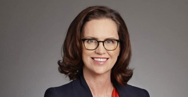 Head and shoulders shot of smiling Shell Australia chair Cecile Wake wearing a dark suit, red shirt, and dark-framed eyeglasses